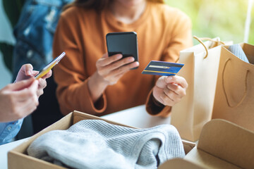 Two women using mobile phone and credit card for online shopping with shopping bag and postal parcel box of clothing on the table