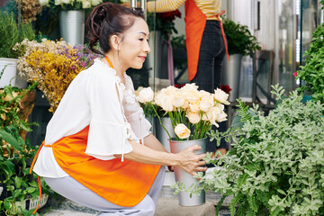 Smiling senior Vietnamese woman arranging buckets of flowers in her shop