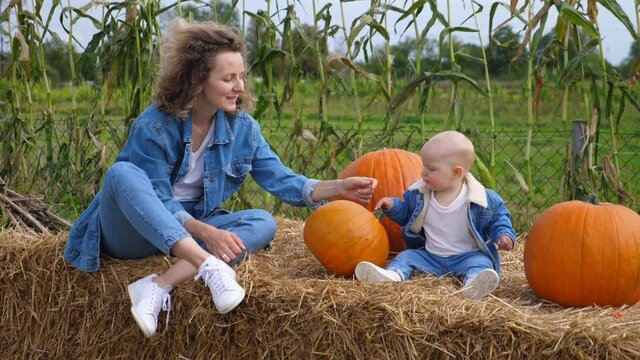 Happy single mom and her baby in a matching denim outfits siting on a hale bale playing with the pumpkins. Mother daughter bond