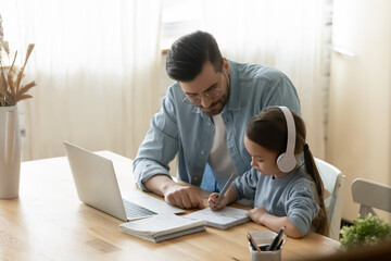 Focused little adorable european girl sitting at desk with wireless headphones, involved in e-learning with caring young father at home. Daddy helping small 7s old kid daughter preparing homework.