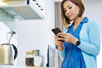 Pretty young Asian woman waiting for dripping coffee on kitchen counter and answering text messages on her smartphone