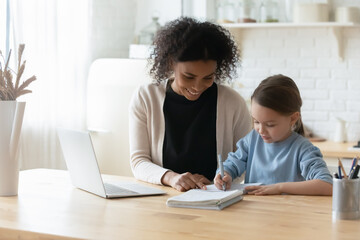 Obraz premium Happy beautiful african ethnicity woman tutor giving private educational lesson to focused small kid girl. Smiling mixed race mother helping little adopted child daughter with school homework.