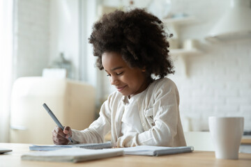 Smiling adorable beautiful african ethnicity little girl sitting at table, writing notes in copybook. Happy small school aged mixed race kid involved in preparing interesting homework alone indoors.