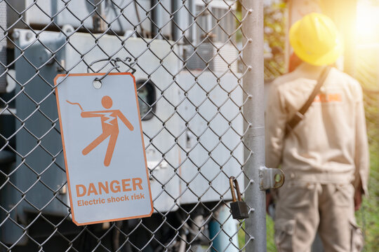 Warning sign of electric shock danger Hanging on a metal mesh fence. 
With an electrician doing maintenance of the equipment inside.