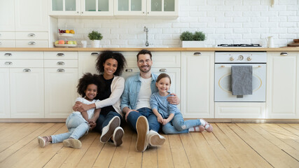 Full length portrait relaxed happy multiracial couple with cute kids sitting on warm wooden floor in modern kitchen. Smiling mixed race family homeowners enjoying peaceful stress free weekend time. - Powered by Adobe
