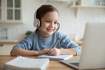 Head shot happy cute little 7s old kid girl wearing earphones, looking in laptop screen, involved in online foreign language private lesson with tutor or enjoying distant e-learning school class.