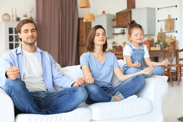 Obraz premium Calm young family with little daughter sit on couch practice yoga together, happy parents with small preschooler girl child rest on sofa meditate relieve negative emotions on weekend at home