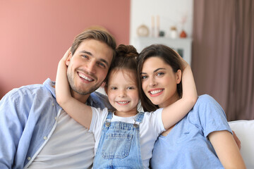 Positive friendly young parents with smiling little daughter sitting on sofa together while...