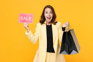Fototapeta premium Surprised young brunette woman 20s wearing basic light suit jacket hold package bags with purchases after shopping sign with SALE title isolated on yellow background, studio portrait. Black friday.