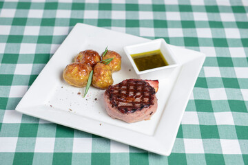 Grilled beefsteak, served with baked potatoes and sauce on a white plate over green plaid tablecloth.