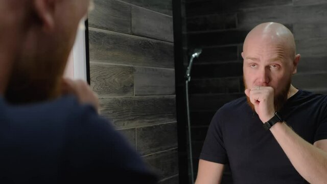 A young, Caucasian, charismatic, bald guy with a red beard yawns in front of the mirror, covering his mouth with his fist. Sleepy reflection. Morning awakening. Close-up.