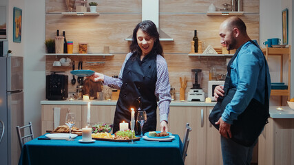 Young caucasian lady cooking for her husband romantic dinner, waiting in kitchen. Woman preparing festive dinner with healty food, man coming back from job surprised kissing and hugging his wife