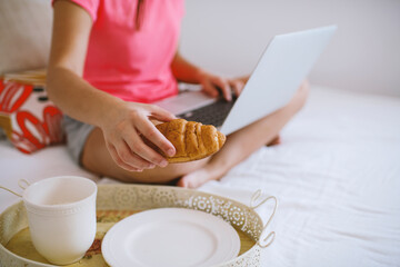 Caucasian girl  sitting on the bed working on a laptop, having breakfast in bed. Work from home, stay home concept, home office, working home, self-isolation.