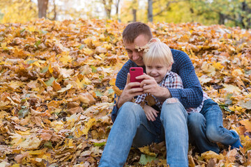 Dad teaches child to take pictures on phone. Father and son sitting in autumn Park.