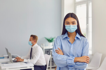 Fototapeta na wymiar Serious young woman wearing face mask standing in her workplace looking at camera