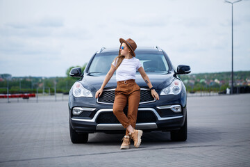 A beautiful girl of European appearance with glasses and a brown hat is standing near a black car....