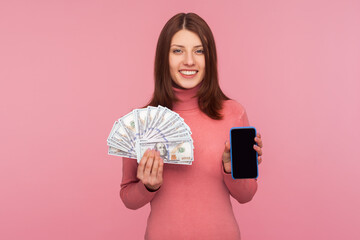 Cheerful brunette woman in pink sweater holding dollars banknotes and cell phone with mock up blank...