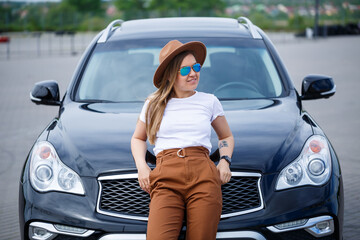 A beautiful girl of European appearance with glasses and a brown hat is standing near a black car. Young woman with car in parking
