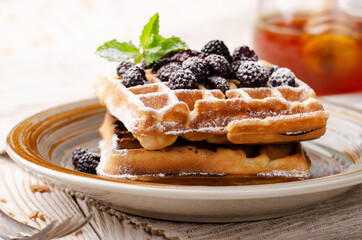 Belgian waffles served with blackerries and mint leaf dusted with powdered sugar on white wooden kitchen table with syrup aside