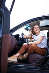 Young beautiful woman with long hair sits in a black car at a parking lot. Pretty girl in casual clothes. Car trip