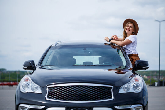 A Beautiful Girl Of European Appearance With Glasses And A Brown Hat Is Standing Near A Black Car. Young Woman With Car In Parking