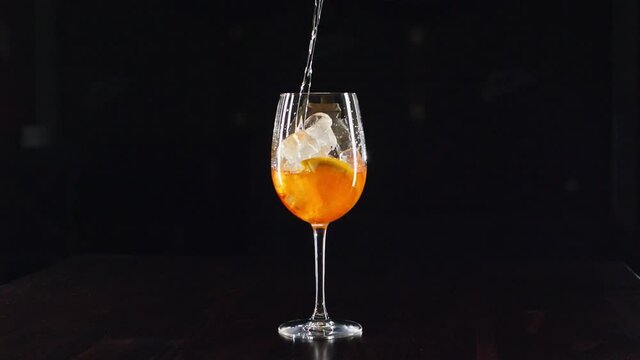 Close up bartenders hands pouring prosecco in wine glass with ice, mixing cocktail with orange juice and alcohol. Long fizzy drink with black background in 4k resolution in slowmotion