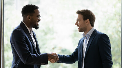 Overjoyed diverse male business partners handshake close deal or make agreement after successful negotiation. Smiling multiracial businessmen shake hands get acquainted greeting in office.