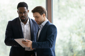 Concentrated multiracial businessmen discuss collaboration use modern tablet in office, focused successful diverse male colleagues brainstorm discuss business project on pad device at meeting.