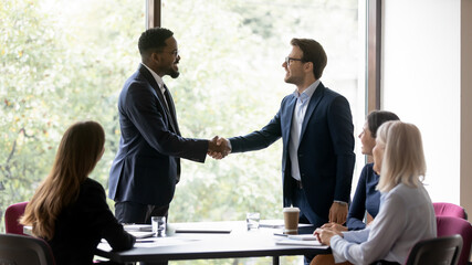 Happy multiracial male business partners handshake close deal or make agreement at office meeting. Smiling diverse businessmen shake hands get acquainted greeting at briefing. Collaboration concept.
