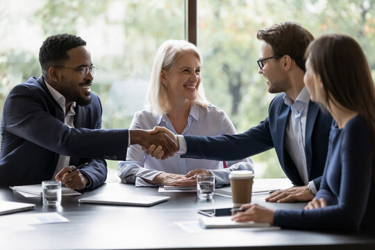 Smiling diverse businesspeople shake hands get acquainted greeting at team meeting in office. Happy multiracial male colleagues employees handshake at briefing with coworkers. Acquaintance concept.