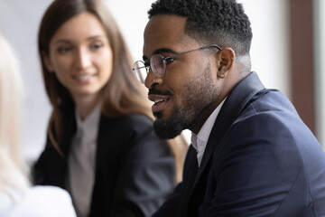 Close up of confident young African American male employee in glasses brainstorm at team meeting with colleagues. Biracial businessman talk discuss idea with multiracial coworkers at office briefing.