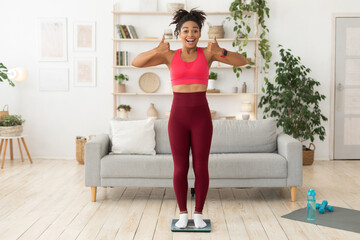 Black Fitness Girl Gesturing Thumbs-Up Standing On Scales At Home