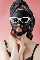 Young woman in glasses appling black cosmetic fabric facial mask on pink background. Face peeling mask with charcoal, spa beauty treatment, skincare, cosmetology. Close up