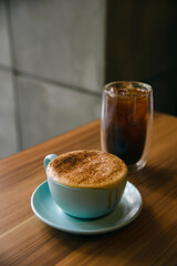 Hot cappuccino in coffee cup on the wooden table with ice americano in background.