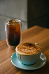 Hot cappuccino in coffee cup on the wooden table with ice amaricano in background.