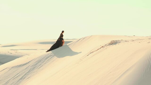 A beautiful young woman in a luxurious black silk dress walks in the desert. The fabric of a long train flutters in the wind in slow motion. Background desert white sand dunes, blue sky