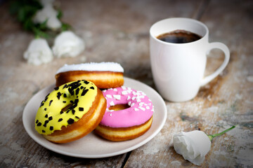 Colored donuts on a plate with a cup of coffee on a wooden table. Light flowers lie nearby