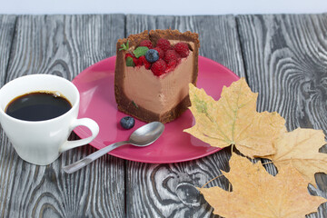 A piece of no-bake chocolate cake with cream cheese. Garnished with raspberries, blueberries and mint leaves. Nearby is a cup of coffee and dried maple leaves.