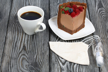 A piece of no-bake chocolate cake with cream cheese. Garnished with raspberries, blueberries and mint leaves. Next to it is a cup of coffee and a medical mask with antiseptic.