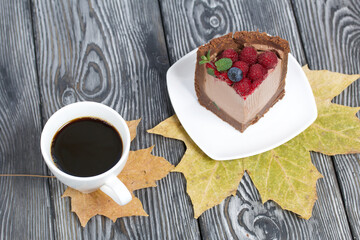 A piece of no-bake chocolate cake with cream cheese. Garnished with raspberries, blueberries and mint leaves. Nearby is a cup of coffee and dried maple leaves.