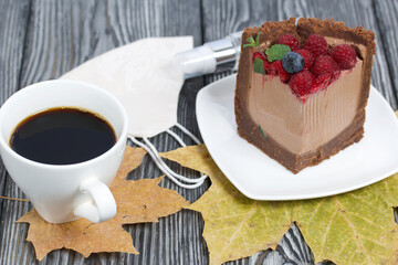 A piece of no-bake chocolate cake with cream cheese. Garnished with raspberries, blueberries and mint leaves. Next to it is a cup of coffee, a medical mask with antiseptic and dried maple leaves.