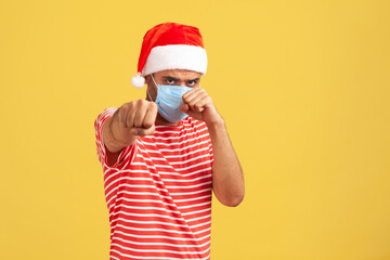 Confident man in santa claus hat with surgical medical mask standing in defensive posture clenched fists, ready to fight with coronavirus covid-19. Indoor studio shot isolated on yellow background