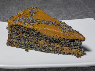 A slice of poppy seed pie. Top of the cake is covered with light brown cream of boiled condensed milk. The pie is on a rectangular white plate