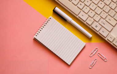 Creative flat lay, photo of workspace desk with a computer keyboard, notepad, a pen, some paper clips, copy space, pink and yellow background, minimal style, selective focus