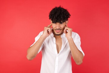 Young arab man with afro hair wearing shirt standing over isolated red background covering ears with fingers with annoyed expression for the noise of loud music. Deaf concept.