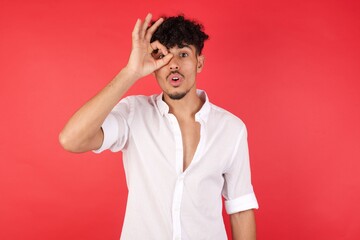 Young arab man with afro hair wearing shirt standing over isolated red background doing ok gesture shocked with surprised face, eye looking through fingers. Unbelieving expression.