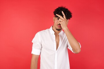Young arab man with afro hair wearing shirt standing over isolated red background peeking in shock covering face and eyes with hand, looking through fingers with embarrassed expression.