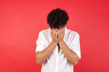 Fototapeta premium Young arab man with afro hair wearing shirt standing over isolated red background covering her face with his hands, being devastated and crying.