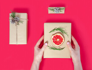 Three gift boxes made of Kraft paper and women's hands on a red background. The concept of zero waste.Eco-friendly natural decor for packaging Christmas and new year gifts.flat lay
