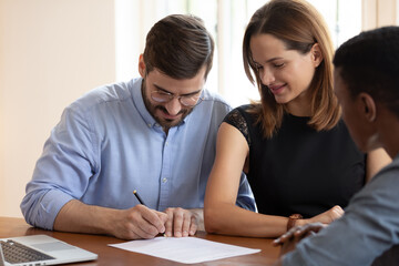 Happy young couple sit at desk meet with realtor put signature on contract buying first house together, excited husband and wife sign business agreement close deal with agent at office meeting